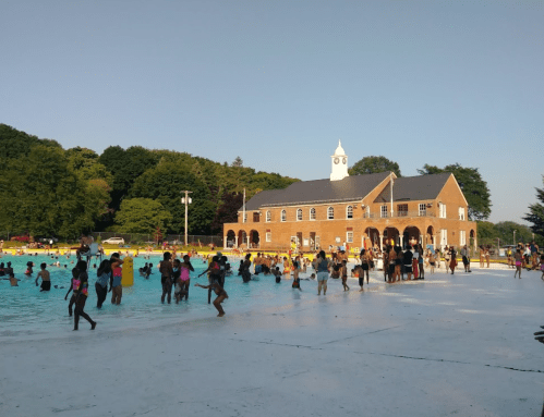 A crowded outdoor pool with people swimming and playing, surrounded by trees and a brick building in the background.