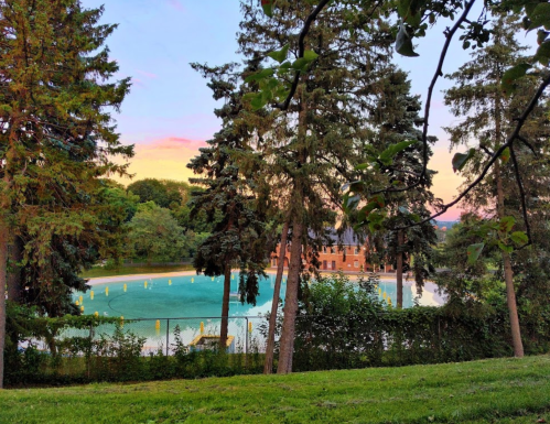 A serene landscape featuring a pond surrounded by trees and a distant building, with a colorful sunset in the background.