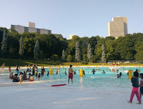 A crowded outdoor pool with people swimming and playing, surrounded by trees and tall buildings in the background.