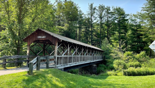 A wooden covered bridge spans a small stream, surrounded by lush green trees under a clear blue sky.