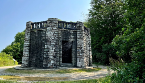 A stone structure with a circular design, surrounded by greenery and a gravel path under a clear blue sky.