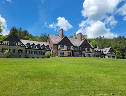 A large, elegant mansion with a mix of stone and wood architecture, set on a green lawn under a blue sky with clouds.