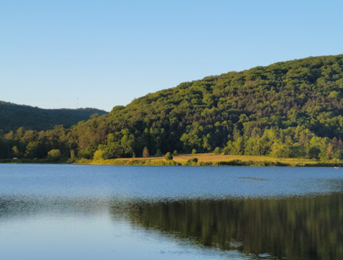 A serene lake surrounded by lush green hills under a clear blue sky, reflecting the landscape in the water.