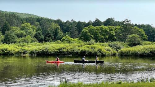 Two boats on a calm river surrounded by lush greenery; one red kayak and one black canoe with three people.
