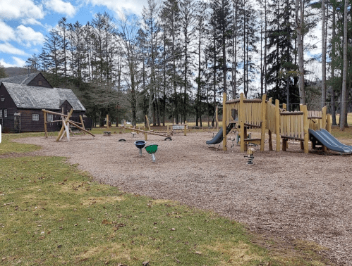 A playground with wooden structures, slides, and swings, surrounded by trees and a grassy area.