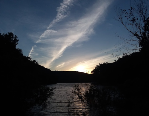 Sunset over a calm river, with silhouettes of trees and hills against a colorful sky.