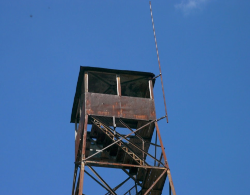 A tall, rusty watchtower against a clear blue sky, featuring a small roof and a single antenna.
