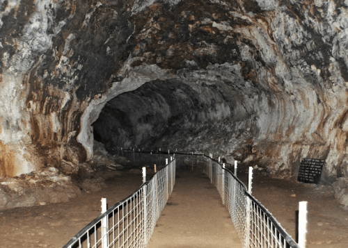 A dimly lit cave interior with a gravel path and metal railing leading into a dark tunnel.