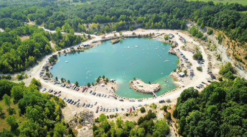 Aerial view of a circular quarry lake surrounded by trees, with parked cars and people enjoying the water.