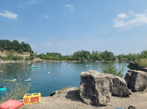 A serene lake scene with people kayaking and relaxing by the water, surrounded by greenery and rocky shores.