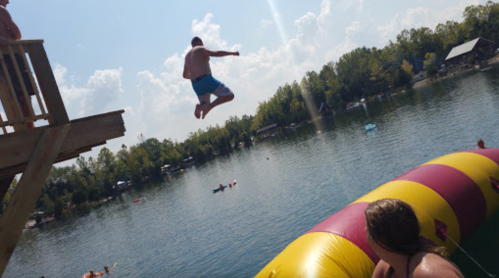 A person jumps off a platform into a lake, aiming for a colorful inflatable blob, with others enjoying the water nearby.