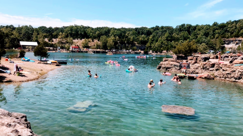 A sunny day at a swimming area with people enjoying the water, surrounded by rocky shores and greenery.