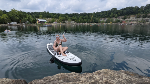 A person paddles on a stand-up paddleboard in calm water, surrounded by trees and a rocky shoreline.