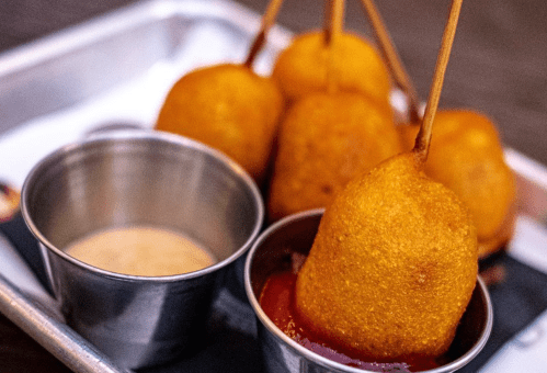 A close-up of golden corn dogs on sticks served with dipping sauces in metal cups.