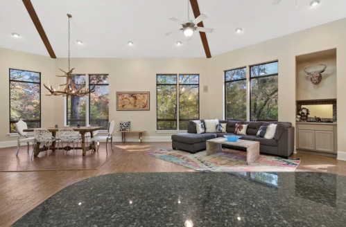 Spacious living area with large windows, modern furniture, and a dining table, featuring natural light and rustic decor.