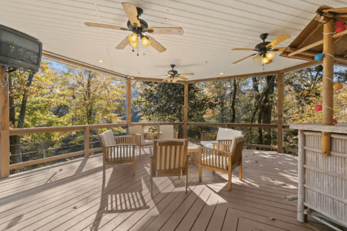 A spacious outdoor deck with wooden furniture, ceiling fans, and a scenic view of trees in autumn.