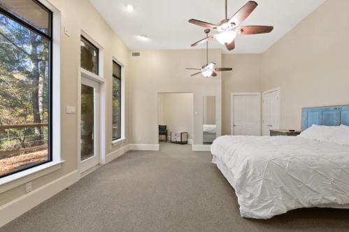 Spacious bedroom with large windows, a ceiling fan, and a light-colored bedspread on a king-size bed.