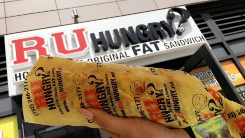 A hand holds a wrapped sandwich in front of the "RU Hungry?" restaurant sign.