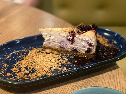 A slice of cheesecake topped with berry sauce and crumbs, served on a blue plate on a wooden table.