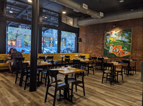 Interior of a restaurant featuring wooden tables, black chairs, and a colorful mural on a brick wall.