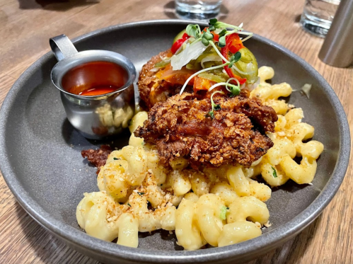 A plate of macaroni and cheese topped with fried chicken, garnished with vegetables and served with a small cup of sauce.