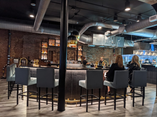 A modern bar with high stools, exposed brick walls, and a sleek design, featuring two women seated at the counter.