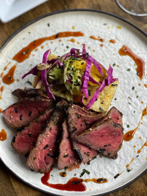 A plate featuring sliced steak, topped with colorful slaw and served on a toasted bread, drizzled with sauce.