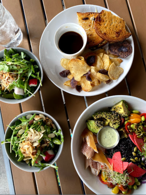 A table with two salads, a plate of chips and toast, and a colorful bowl of fruit and vegetables with dressing.