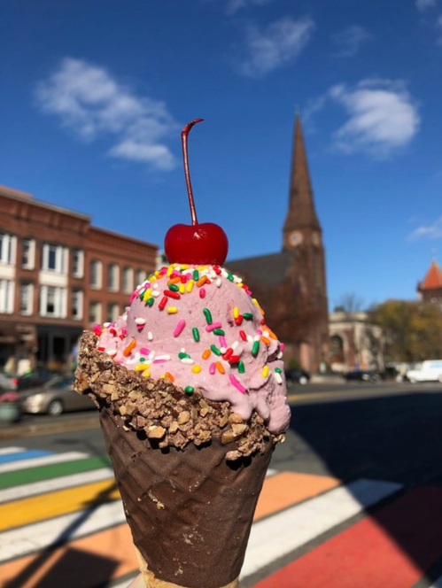 A close-up of a colorful ice cream cone topped with a cherry, with a church steeple and blue sky in the background.