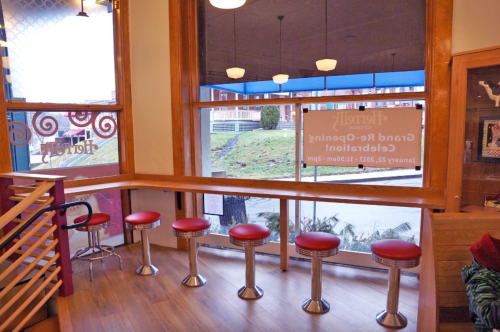 Interior of a café with red stools at a wooden counter, large windows, and a sign for a grand reopening event.