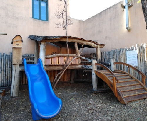 A wooden play structure with a slide and bridge, surrounded by a fence and a tree, in a sunny outdoor area.
