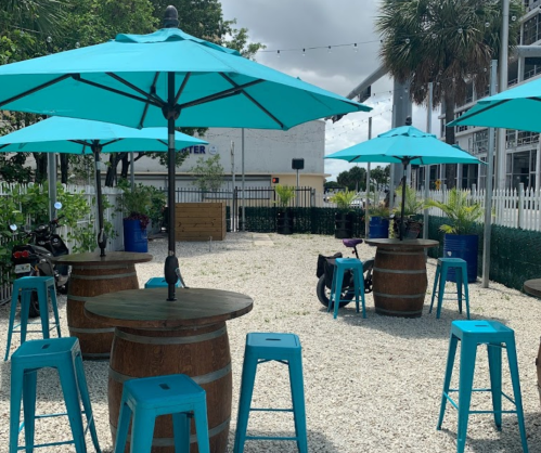 Outdoor seating area with turquoise umbrellas, wooden tables, and barrels on a gravel surface, surrounded by greenery.