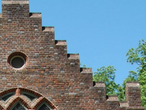Close-up of a brick building's gable with a circular window and greenery in the background against a clear blue sky.