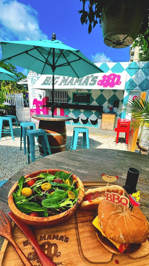 A vibrant outdoor dining area featuring a salad and BBQ sandwich, with colorful tables and a food stall in the background.