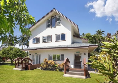 A modern two-story house surrounded by greenery and palm trees under a clear blue sky.