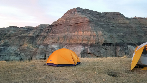 Two orange tents set up on grassy ground with a rocky, layered cliff in the background during sunset.