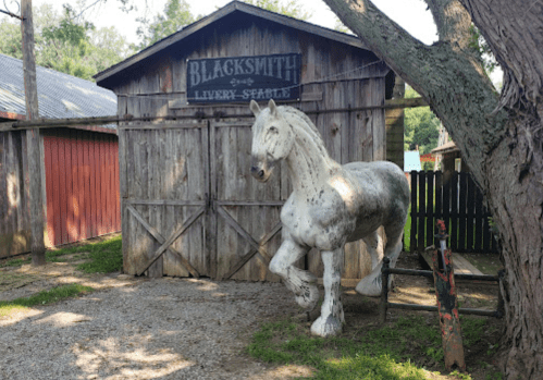 A white horse statue stands in front of a rustic blacksmith barn surrounded by trees and gravel.