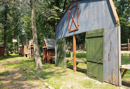 A row of rustic wooden structures with green and blue exteriors, surrounded by trees in a sunny outdoor setting.