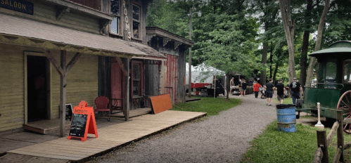 A rustic saloon with a wooden porch, surrounded by trees and a path leading to a gathering of people in the distance.