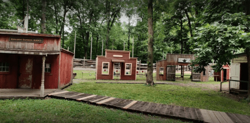 A rustic outdoor scene featuring wooden buildings in a forested area, with a pathway leading through the greenery.