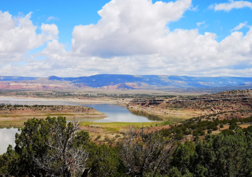 A scenic view of a lake surrounded by hills and clouds under a bright blue sky.