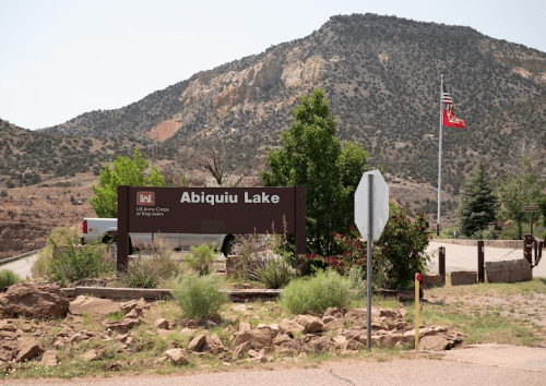 Sign for Abiquiu Lake with mountains in the background and flags nearby, surrounded by greenery and rocky terrain.