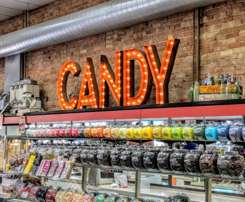 Brightly lit "CANDY" sign above a colorful display of jars filled with various candies in a vintage shop.