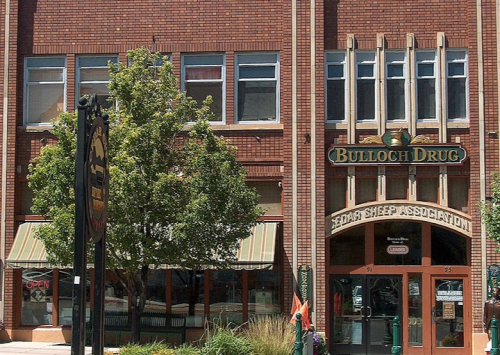 Historic brick building featuring "Bulloch Drug" and "Sedgwick Sheep Association" signs, with trees and outdoor seating.