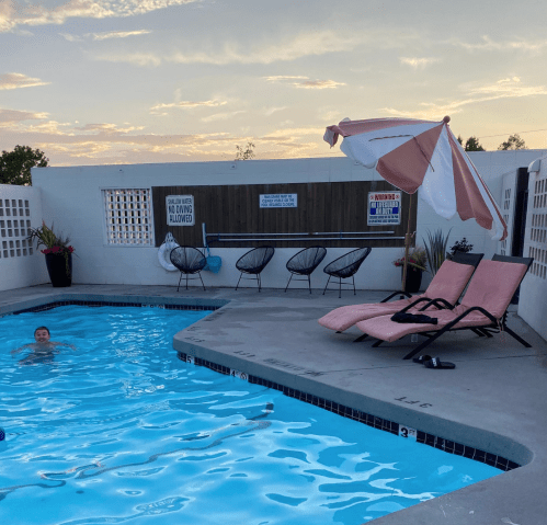 A serene pool scene at sunset, featuring a swimmer, lounge chairs, and a pink umbrella.