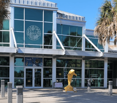 A modern building with large glass windows and a golden sculpture in front, surrounded by palm trees.