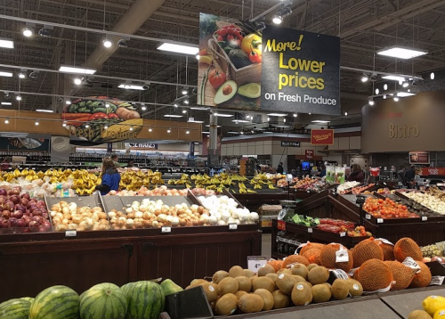 A grocery store produce section filled with various fruits and vegetables, featuring a sign about lower prices.