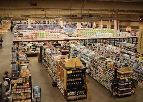 A wide view of a grocery store aisle filled with various products on shelves, with shoppers in the background.