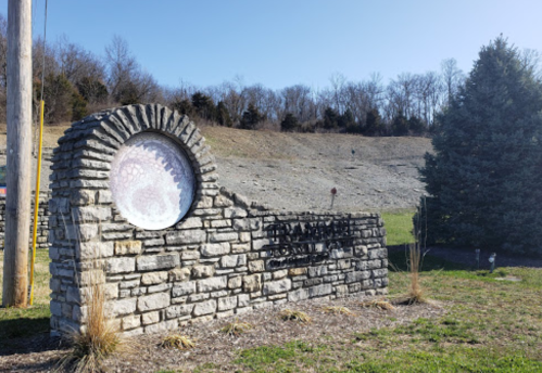 A stone monument with a circular feature, set against a grassy area and trees in the background under a clear sky.