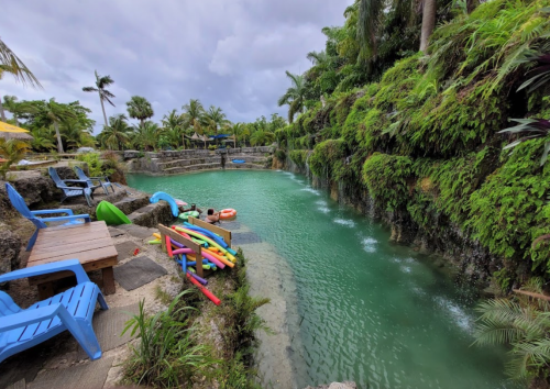 A serene lagoon surrounded by lush greenery, with colorful kayaks and lounge chairs nearby under a cloudy sky.
